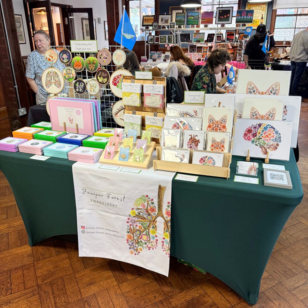 Photo of a market stall featuring embroidery kits and art prints. The banner reads Juniper Forest Embroidery. The tablecloth is a dark green. The floor is wooden. There are other stalls behind. The stall set up is very colourful. 
