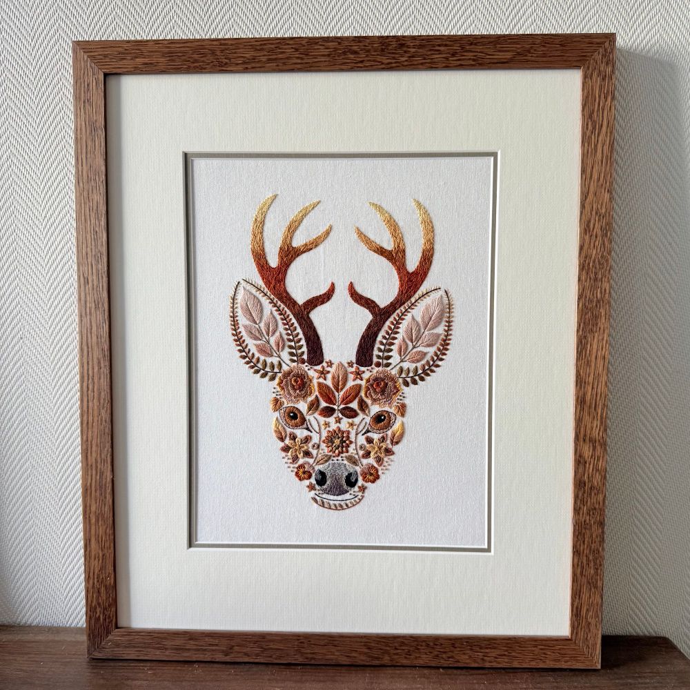 Close-up of a framed embroidered stag’s head artwork. The antlers are shaded from dark brown to golden yellow, and the face is formed from intricate floral and leaf embroidery in autumnal tones. The piece is set in a wooden frame with a cream mount.