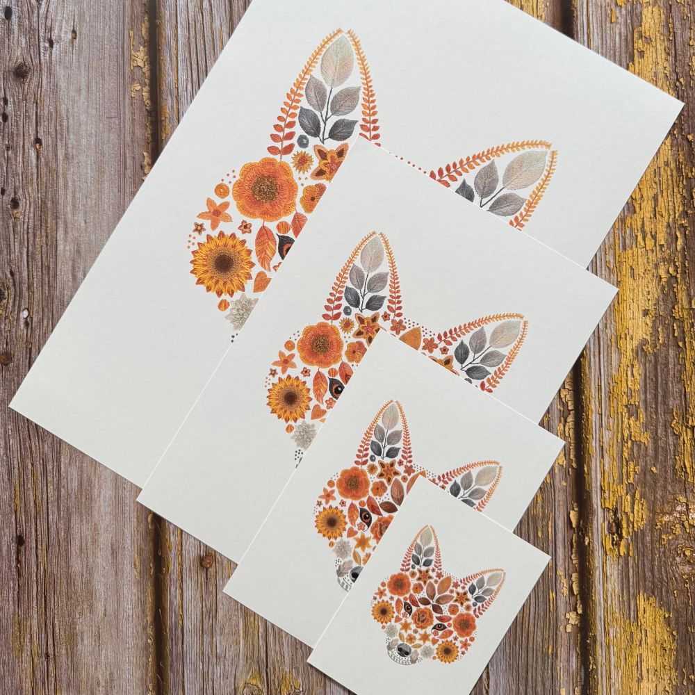 A photo showing 4 floral fox art prints. The art prints are of the same image but are in 4 different sizes. The design is of a foxes face created from flowers and leaves. The largest print is at the back with the smallest at the front of the stack of art prints. They are displayed diagonally. The background is brown wood. 