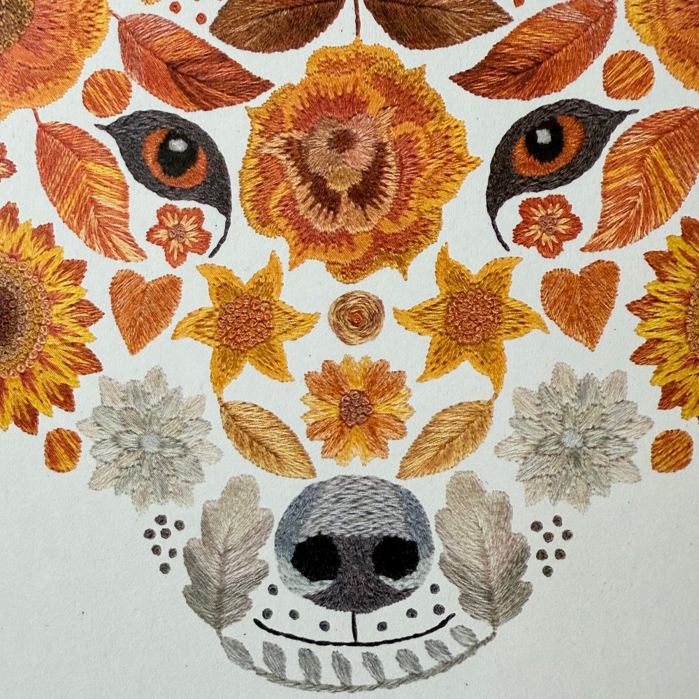 Close up of the foxes face on the floral fox art print. You can see the eyes, nose and mouth. The embroidery stitches are visible and show lots of texture. The colours are various shades of orange and various shades of grey. 