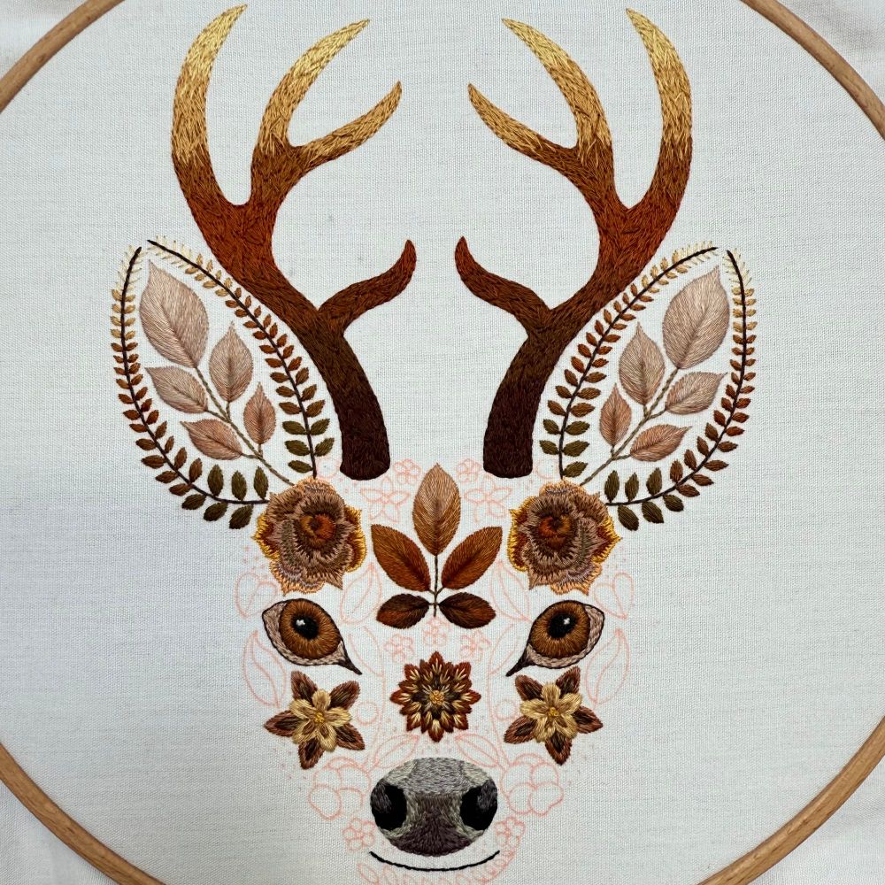 Photo of an embroidery of a stags face. The face is created using flowers and leaves. The photo shows the face, ears and antlers. The areas of embroidery are created using various brown threads. The areas yet to be embroidered are marked out in red fabric pen. The fabric is white. 