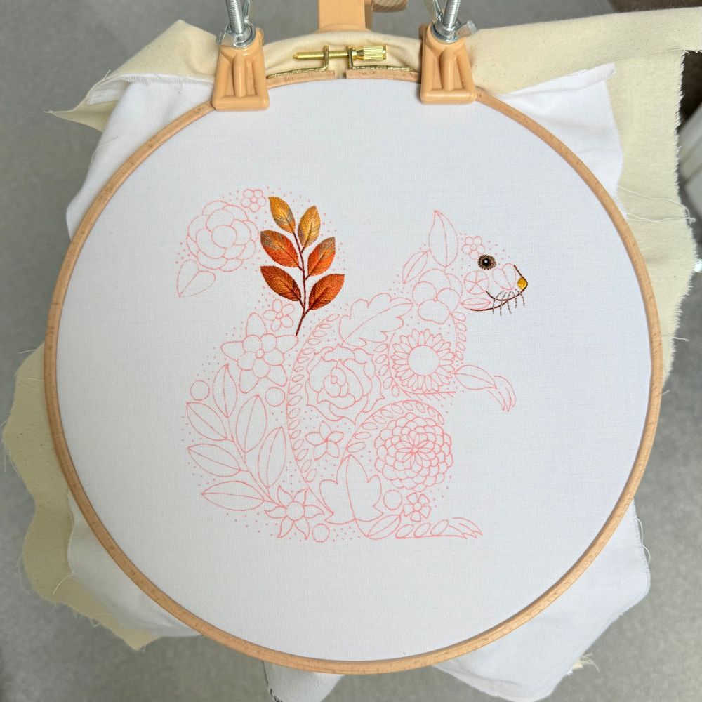 A full view of an embroidery hoop containing a red squirrel pattern made up of floral elements. The squirrel's face and a leaf cluster on its tail are fully stitched in autumn tones, while the rest of the design is outlined in pink fabric pen, awaiting embroidery.