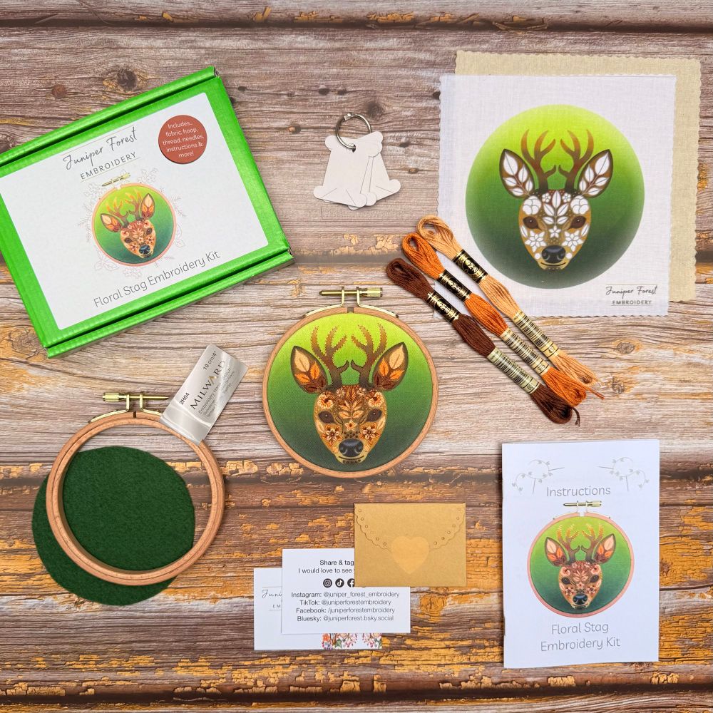 Floral Stag Embroidery Kit contents—printed fabric, wooden hoop, skeins of brown embroidery thread, needles, instruction booklet and packaging arranged on wood surface