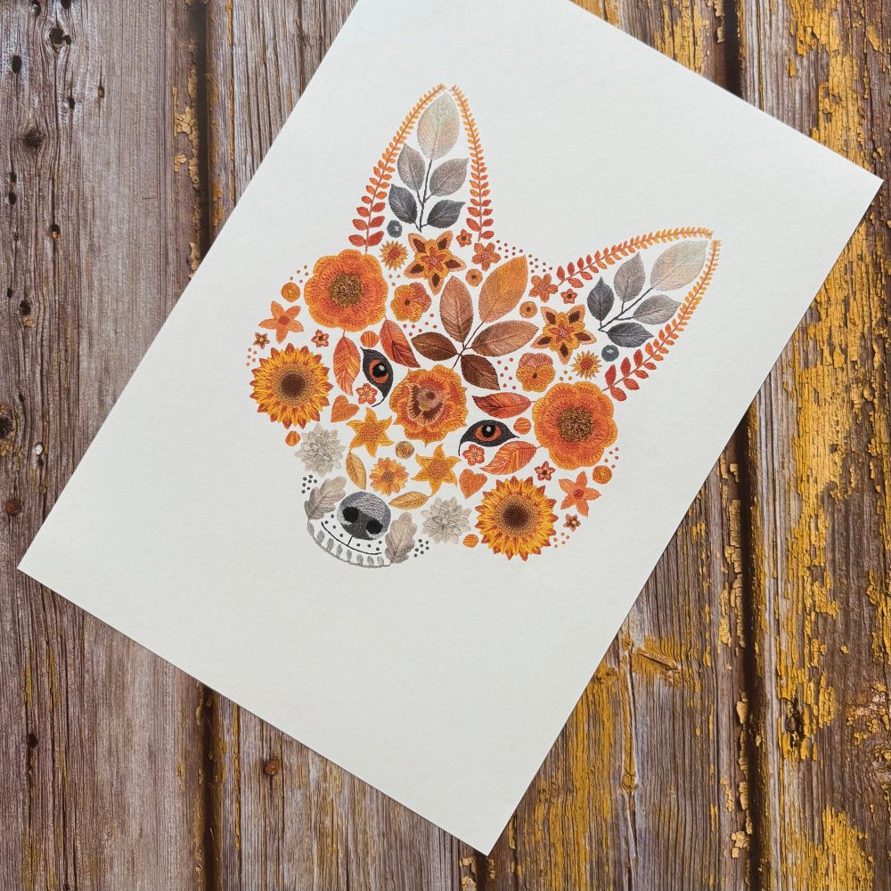 A photo showing a floral fox art print. The design is of a foxes face created from flowers and leaves. The art print is displayed diagonally. The background is brown wood. 