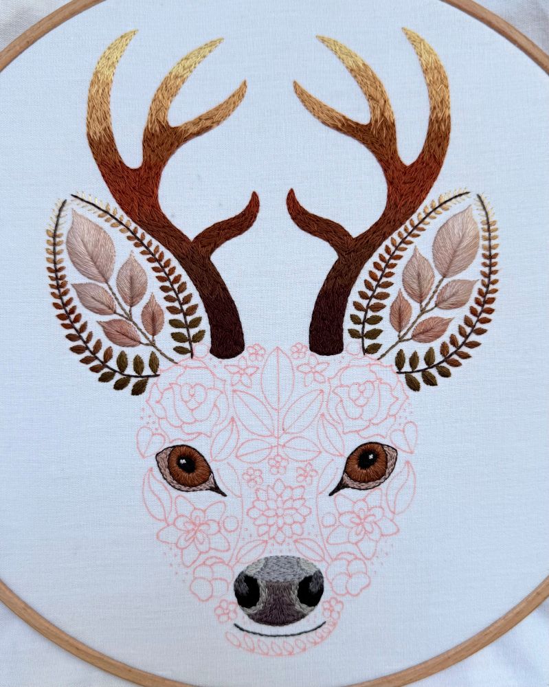 Photo of an embroidery of a floral stag design. The face of the stag is created using flowers and leaves. So far the eyes, nose, mouth, ears and antlers have been stitched in various shades of brown. The face of the stag has been drawn out in red fabric pen. The stag embroidery is on white fabric. 