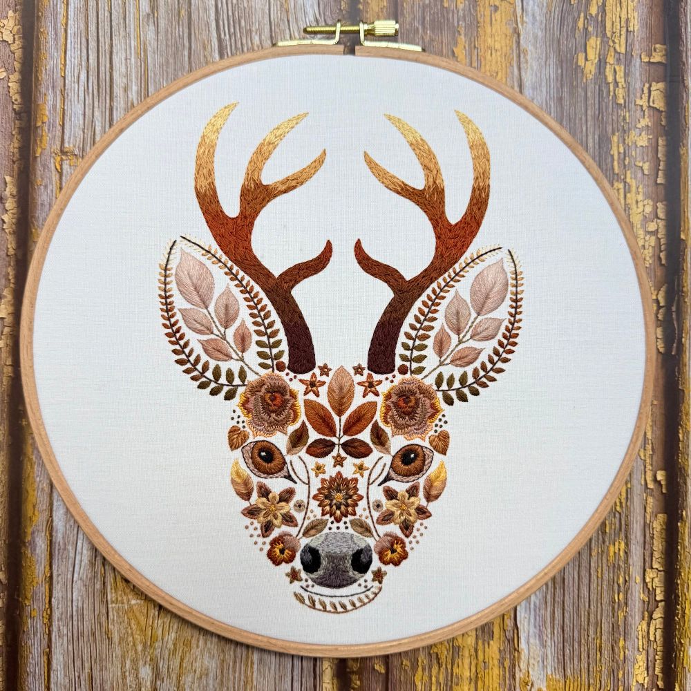 Hand-stitched embroidery of a stag’s head, displayed in a wooden hoop against a rustic wooden background. The design is highly stylized and whimsical, with the entire stag’s face and antlers composed of intricately embroidered flowers, leaves, and botanical elements in warm autumnal shades of brown, gold, rust, and cream. The antlers are shaded from dark to light brown, and the ears are made of leafy motifs. The eyes are created using layered threadwork with floral details surrounding them, giving the piece a magical, nature-inspired appearance.