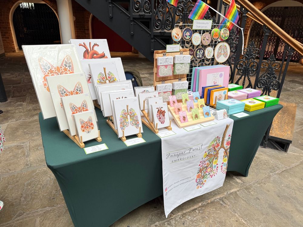 Photo of a craft market stall set up. The table has a dark green tablecloth with a white banner saying Juniper Forest Embroidery. There are art prints displayed on the left side of the table including a floral fox design as well as floral lungs and floral heart. The middle of the table has a wooden display stand with enamel pins. The right side of the table has embroidery kits. The boxes of the kits are brightly coloured including green, yellow, blue, purple, orange and pink. There is a display at the back of the stall showing embroidery hoops. 