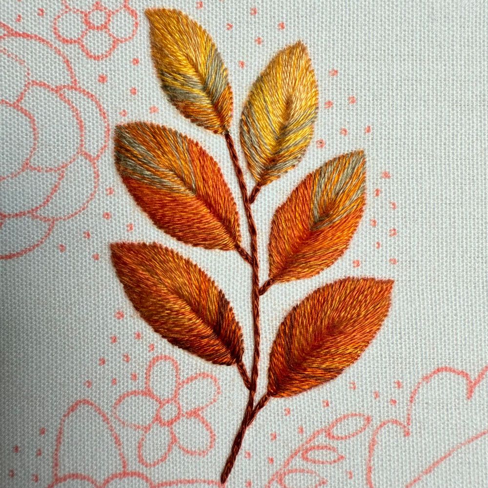 Close-up view of a hand-embroidered leaf cluster in warm autumn tones of orange, gold, and muted gray on white fabric. The stitching is done with leaf stitch, and pink floral guidelines are visible in the background.
