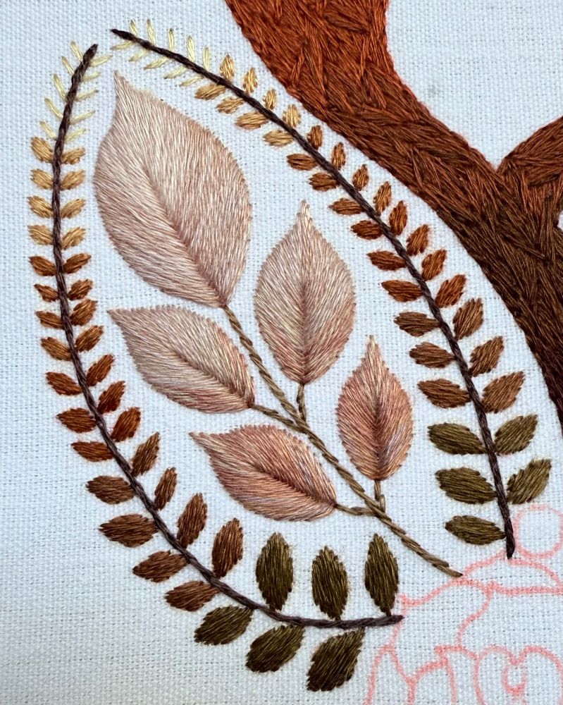 Close up photo of some embroidery. The centre of the embroidery is a cluster of leaves in various shades of pink. Surrounding the leaves are two curved vines. The leaves of the vines have an ombré effect with lighter shades at the top and darker shades at the bottom. The embroidery is stitched on white fabric. 