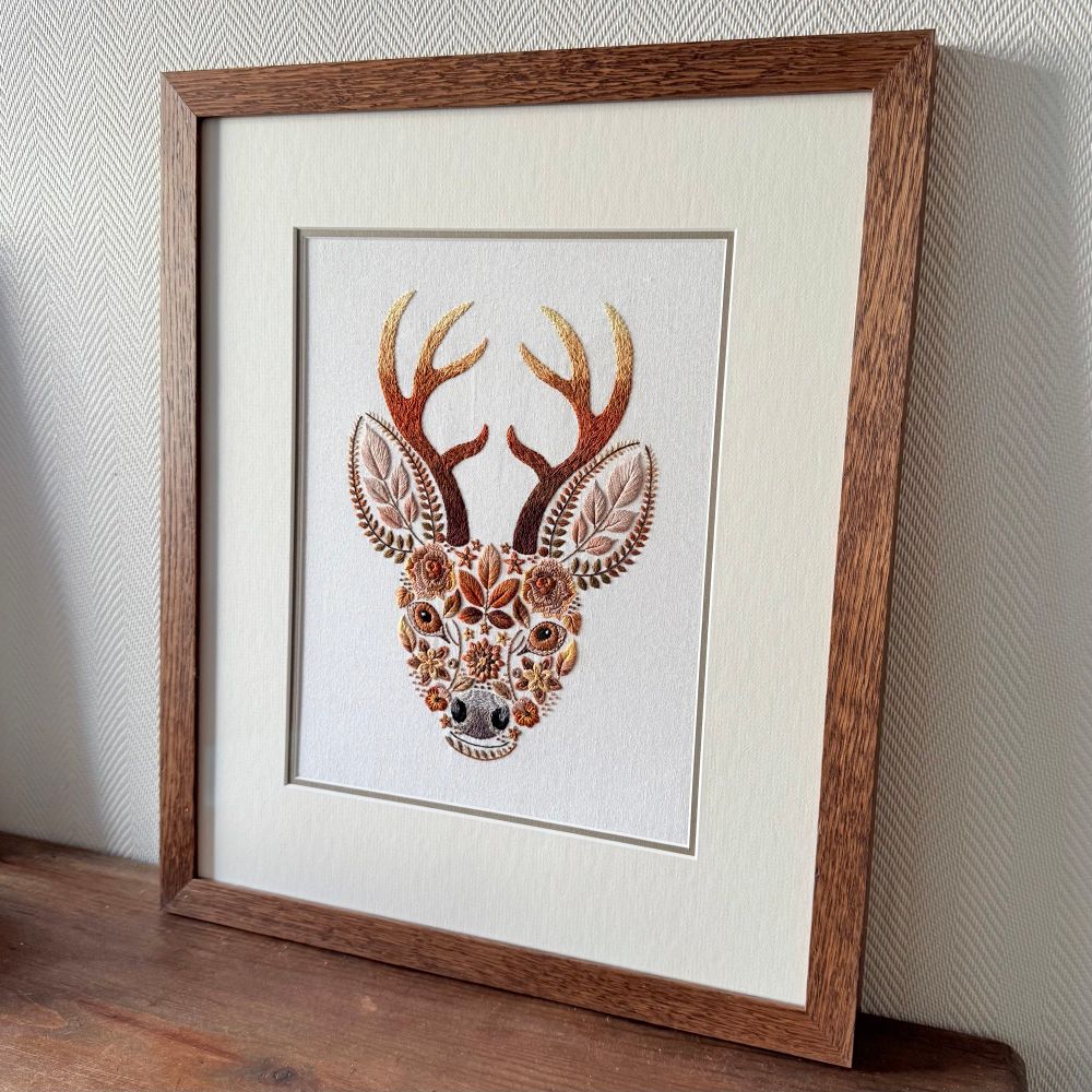 Angled view of the framed embroidered stag’s head artwork, showing the texture and stitching detail. The wooden frame and cream mount surround the richly colored floral and leaf design.