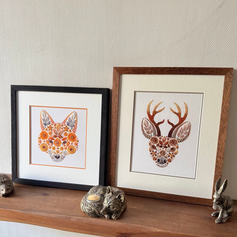 Two framed embroidered artworks side by side on a wooden shelf. The left features a fox’s head in vibrant orange and yellow floral and leaf patterns with a black frame, while the right displays a stag’s head with brown and gold antlers in a wooden frame. Decorative animal figurines, including a hedgehog, sleeping fox candle holder, and hare are placed along the shelf.