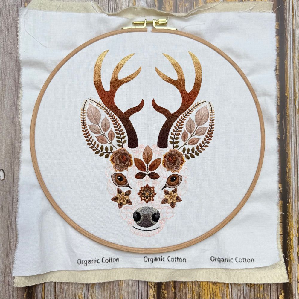 Photo of an embroidery of a stags face. The face is created using flowers and leaves. The photo shows the design in a wooden embroidery hoop. The areas of embroidery are created using brown threads. The areas yet to be embroidered are marked out in red fabric pen. The fabric is white with the words Organic Cotton printed at the bottom of the fabric. The embroidery is shown again a wooden background. 