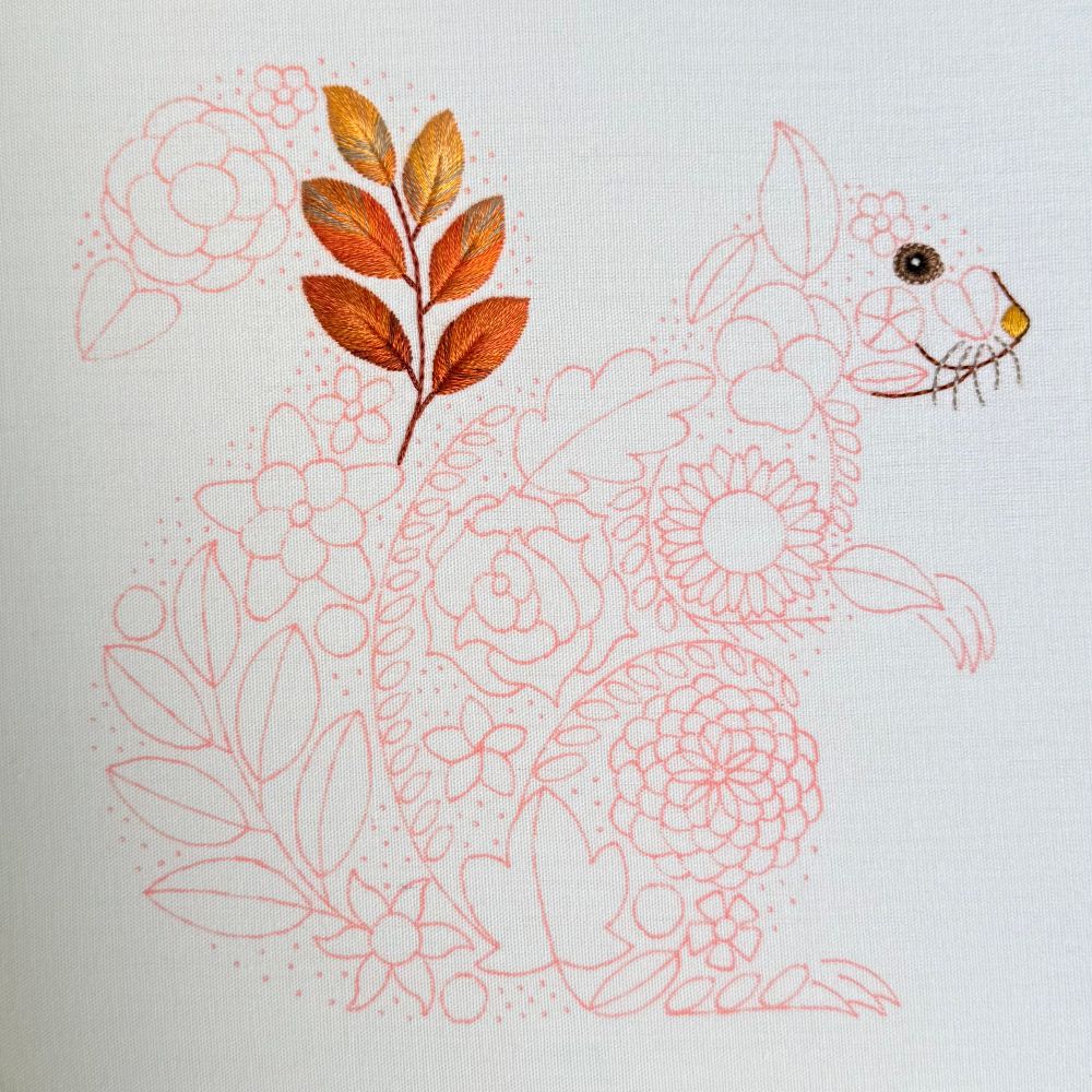 Embroidery in progress showing part of a red squirrel outline filled with floral patterns. The face has been stitched, and a completed cluster of colorful embroidered leaves forms part of the tail. The rest of the design is outlined in pink fabric pen on white fabric.