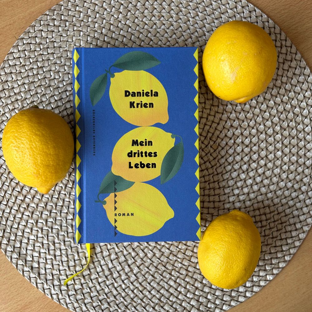 Es ist das Buch „Mein drittes Leben“ von Daniela Krien zu sehen, allerdings in der Ausgabe der Büchergilde Gutenberg. Dort sind auf dem Cover drei Zitronen auf einem blauen Hintergrund abgebildet. Das Buch liegt zusammen mit drei Zitronen auf einem runden Untersetzer. 
