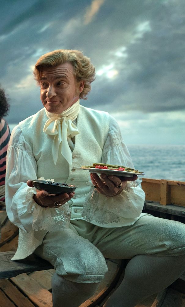 A picture of Stede Bonnet, who is a white blonde man in a white cravat, white vest, and white trousers. He is holding two plates of tapas while in a small boat. 