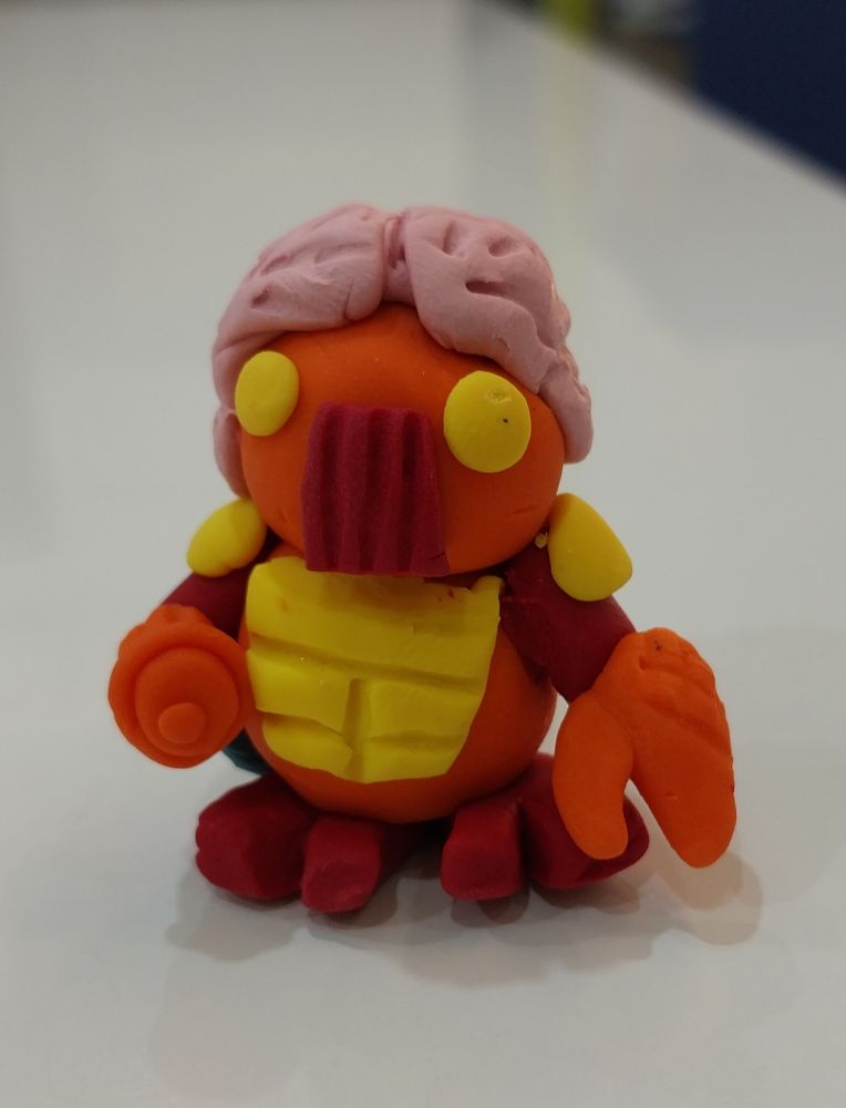 A polymer clay version of Membros ftom the Micronauts; an orange alien with a gun arm and an exposed brain.