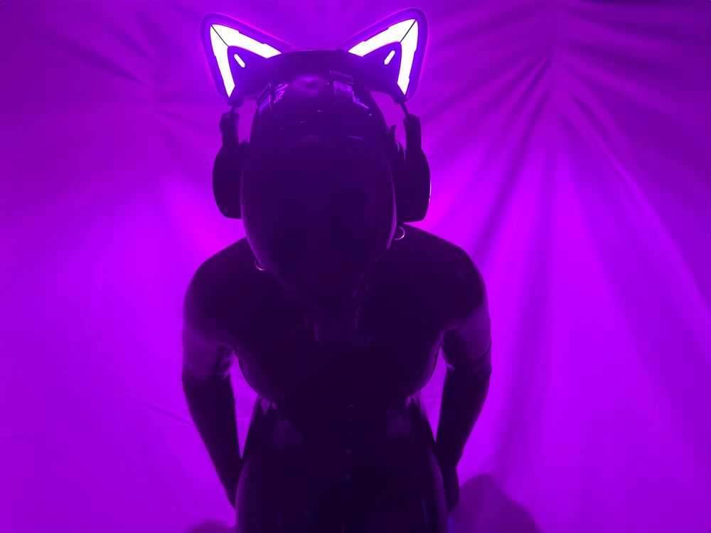 A faceless rubber drone in a neck entry catsuit, MSA millennium gasmask and “control headset” against a light purple lit background looking down at the camera. 