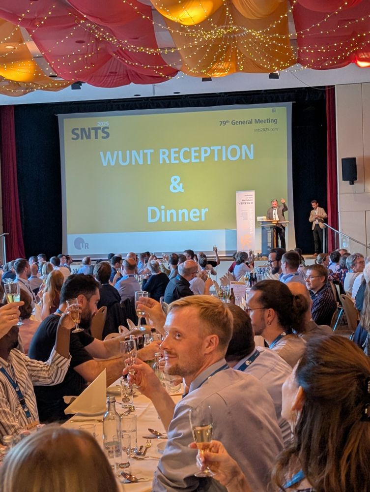 Jörg Frey Standing on the stage of the Kolpinghaus Regensburg, raising a glass, together with a room full of People, cheering to 75 years of WUNT. 