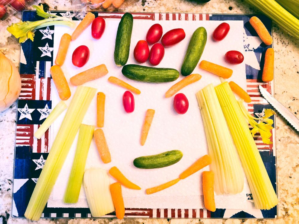 Johnny Joestar's face made out of baby tomatoes, celery, baby carrots, and tiny cucumbers arranged on a 4th of July cutting board and napkin.