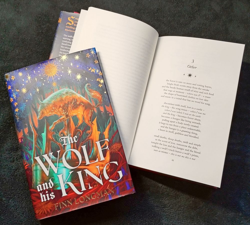 A copy of The Wolf and His King by Finn Longman, open to the first page of chapter 3, which is in verse. Another copy holds the book open.