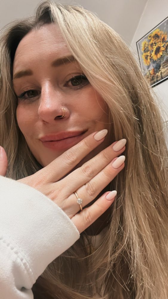Beka, a white woman with blonde hair, wearing a white sweater, posing with her hand covering her chin so you can see her diamond engagement ring and smiling 