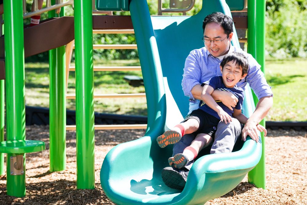 Asian man sliding down a playground slide with young child
