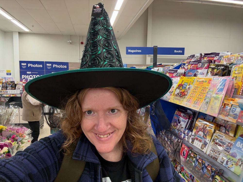 Selfie of a woman with long red hair wearing a green witches hat with black rosette swirls giving an evil grin to camera 