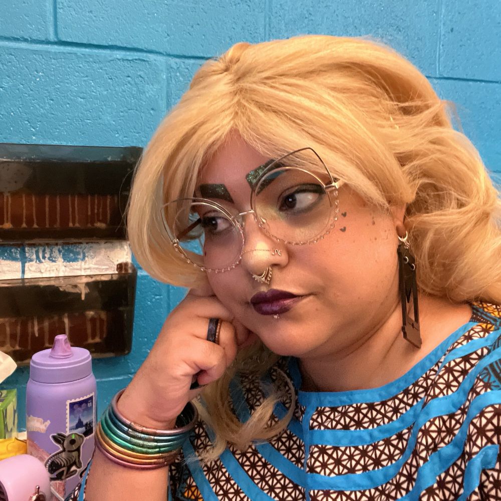 selfie of me! a Black, blonde femme with cats eye shaped wireframe glasses & makeup, looking mildly pensive away from the camera