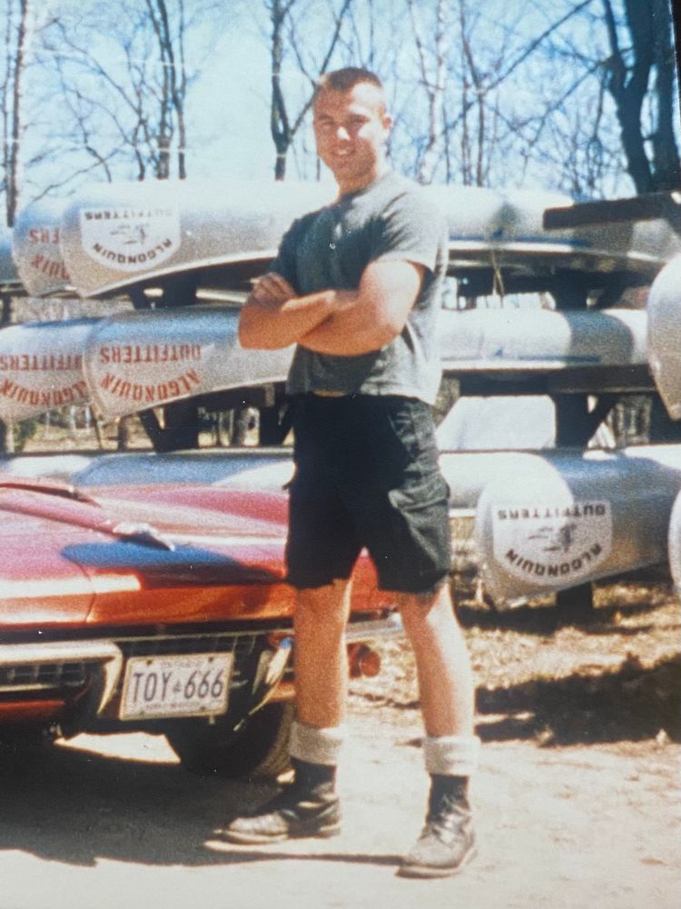 Late teens in cutoff surplus shorts, wool socks, infantry boots, and a t-shirt, feeling like I was cooler than school, posing, arms crossed in front of a 66 Vette and a rack of canoes at the outfitters. Either heading into or coming out of Algonquin Park on a 5 or 6 day canoe trip. 
