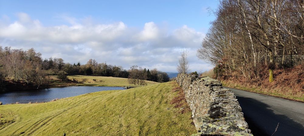 Road at Ghyll Head, dry stone wall, field and tarn in sunshine.