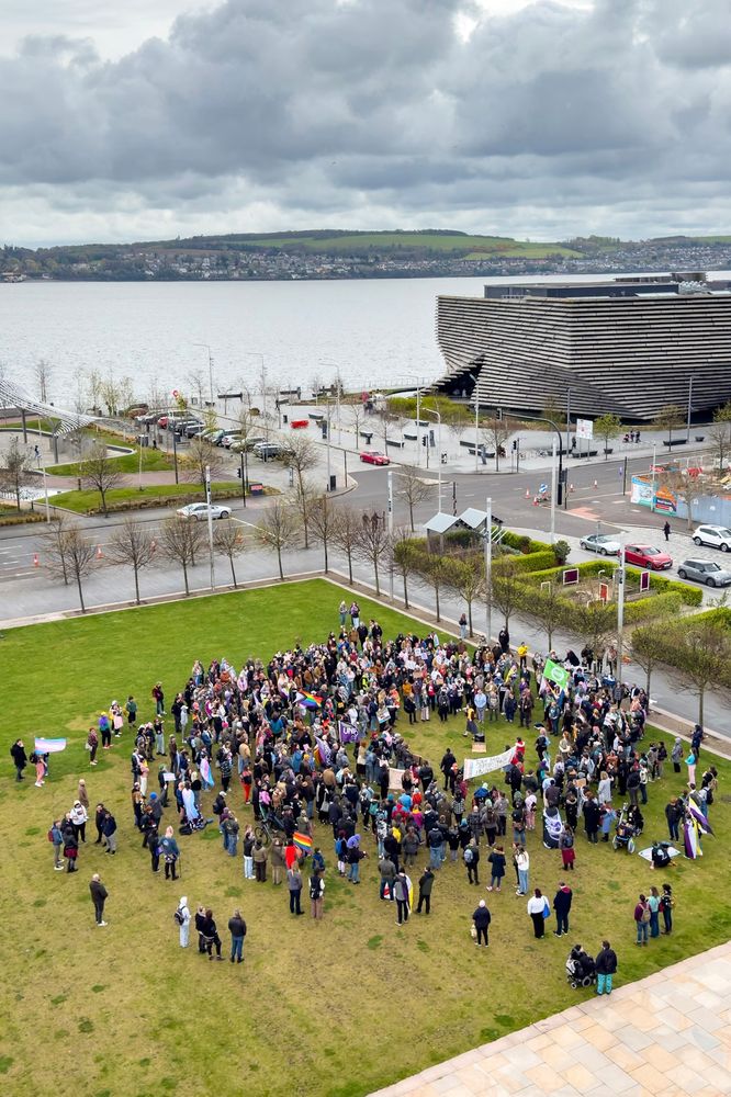 A crowd of perhaps a couple of hundred people, many with trans and other queer flags, gathered in an open grass square beside the river Tay