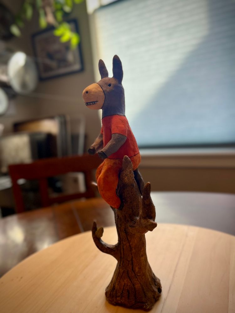 A small smiling donkey sits high atop a barren tree, wearing a matching pair of bright orange pants & a t-shirt.

“Donkey in a Tree” by Sara Swink.