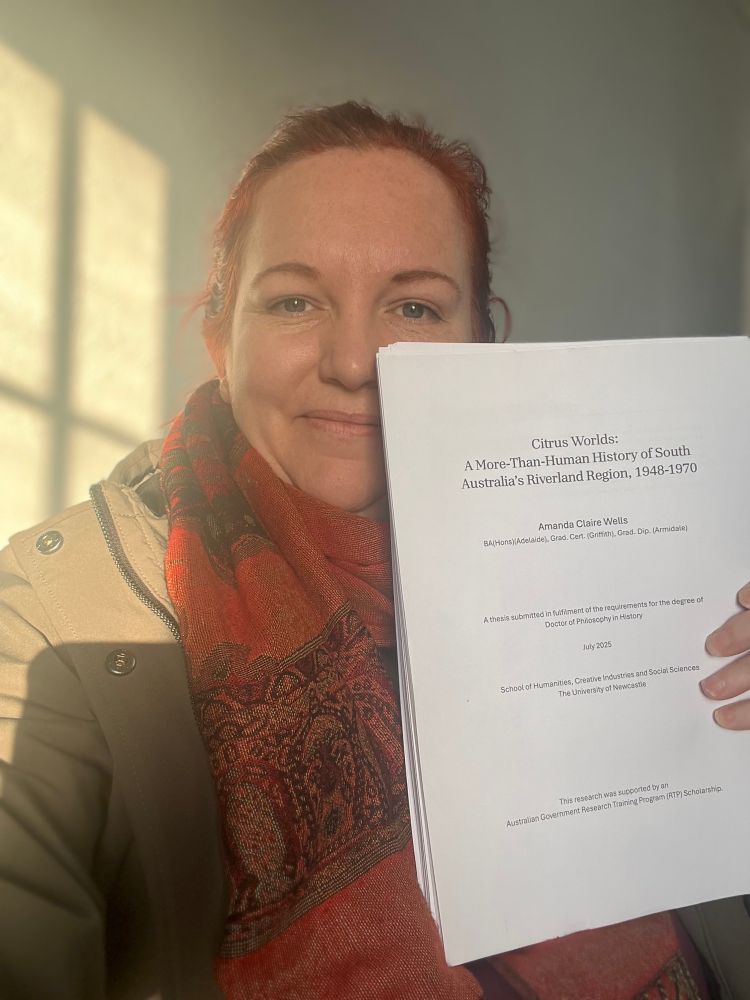 Amanda holding (a chunk of) her PhD thesis, looking happy and relieved, as the sun casts warm light on her right shoulder.