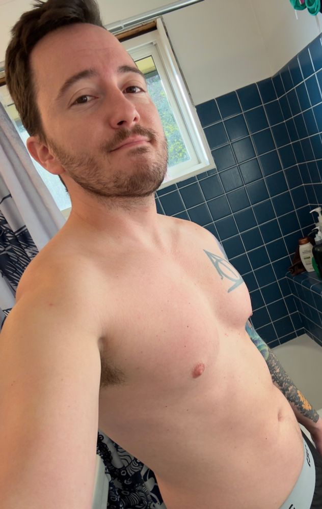 Josh standing shirtless in front of a blue tiled shower. The chest is pumped from doing push ups. 