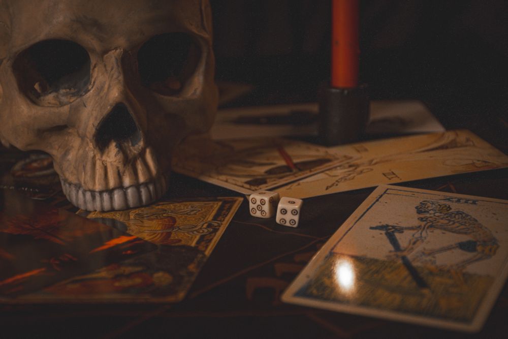 A photo of a skull with no lower jaw set on several spread out tarot cards, they are all the death card. There is a red candle in the photo but the flame cannot be seen at the top of the candle, instead it is visible in one of the death cards where it looks like the image of death is about to reap the flame with his scythe. A pair of bone dice are in the middle with the one pips facing up.