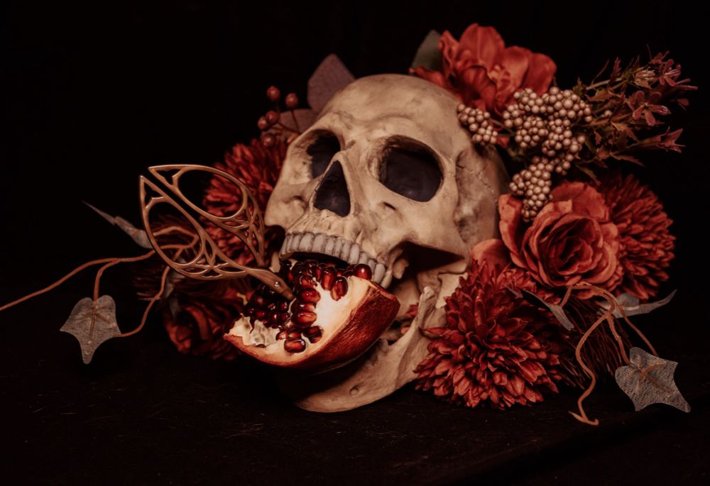 Photo of a skull surrounded by a mixture of muted red flowers. A pomegranate is shoved between the jaws of the skull, and a pair of embroidery scissors are stabbed into the pomegranate. All on a black background. 