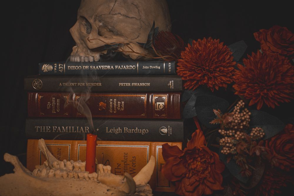 A photo of a skull facing left with no lower jaw on top of a stack of books, a bundle of muted red flowers are to the right of the stack. A hog’s jaw is in front of the book stack facing right, and in the middle of the jaw is a red candle with its flame extinguished and smoke drifting off of the wick. 