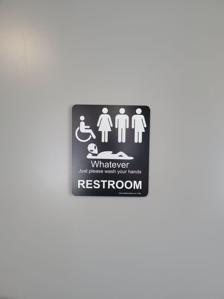 A bathroom sign that says whatever just please wash your hands restroom. It shows a wheelchair user icon, a woman icon, a man icon, a nonbinary icon (wearing half a skirt or dress), and an alien laying down icon