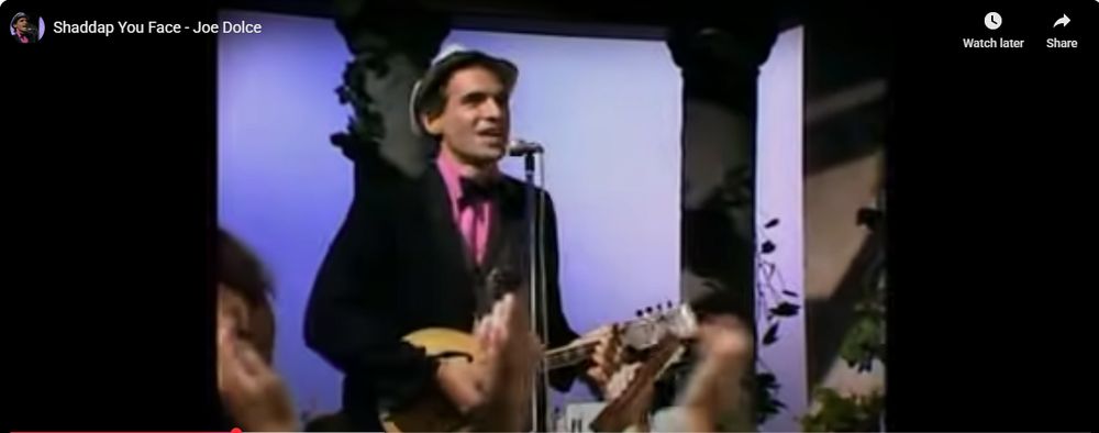 faux-Italian man dressed in a pink shirt with black blazer wearing a small hat playing a guitar. text up top reads "Shaddup you face - Joe Dulce"