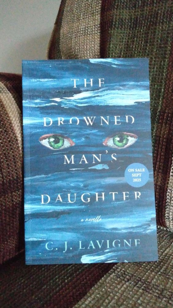 A hardcopy of C. J. Lavigne's The Drowned Man's Daughter, nestled into a couch corner because the cat was not available to do his duty and pose. The cover has green eyes superimposed over a painted ocean. This is an ARC (an advance copy/uncorrected proof), and the cover is stamped "on sale Sept 2025."