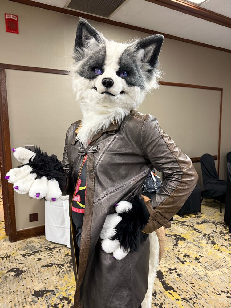 A platinum fox fursuiter wearing a brown leather duster poses in a hotel meeting room