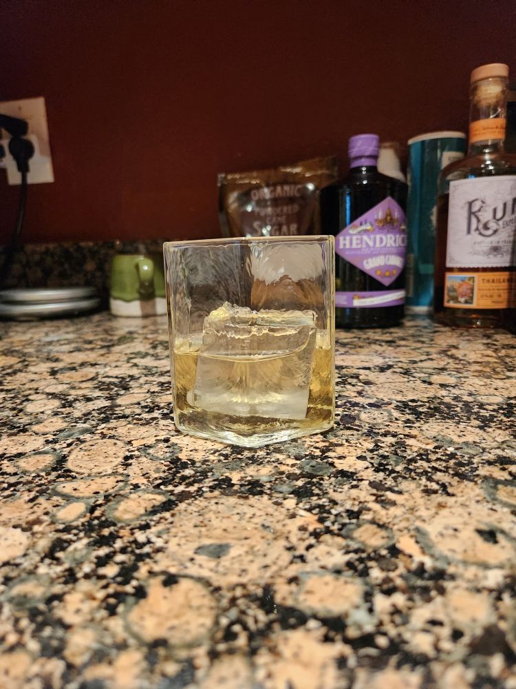 A cocktail glass filled with a nearly clear beverage and large rock of ice