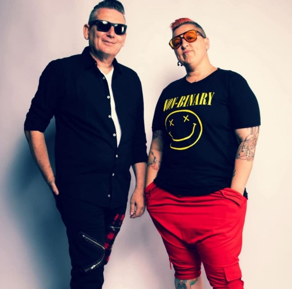 

This is a shot from about the calves up, with ‘The Hardman’ on the left and InkBits on the right.  They are both white skinned humans.  They are standing next to each other, turned slightly inwards. 

The Hardman has short dark, greying hair, he is wearing a white top with a black shirt on top, and black and tartan pants with zippers in them.  He has  dark rayban style sunglasses. He has his left hand in his pocket and the other arm by this side, he is smiling at the camera.
InkBits has short dark hair,  with red colour on top. They are wearing dark framed glasses with yellow lenses and a black t-shirt with a yellow design.  They have red pants that end just past their knees.  Their hands are in their pockets and their head turned slightly back. They have a number of tattoos on their legs and arms and they have an enquiring expression on their face. 
    