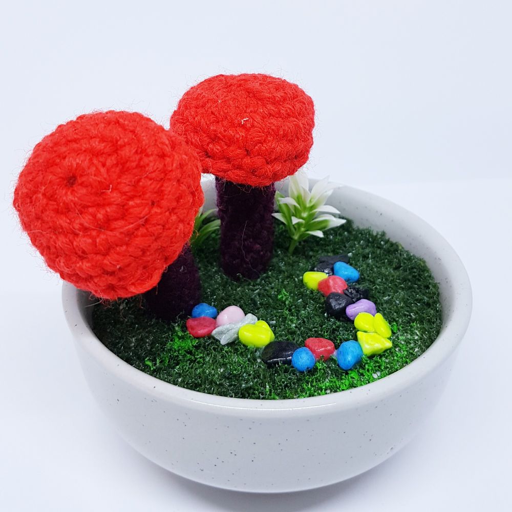 Two red and burgandy, crocheted mushrooms sitting in a little white pot, with fake grass and a little path made from tiny, brightly coloured pebbles . With a little green plant in there with them. 