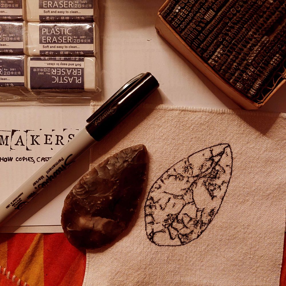 A close cropped image in warm light shows a black ink impression of a small worked flint. Next to it sits the replica the print was made from, and surrounding them both are the remnants of zine making, printed letters, pens, paper and glue