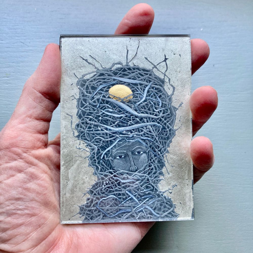 Miniature engraved glass picture of a head wrapped in a birds nest. A gold egg sits on top of the bnest.
