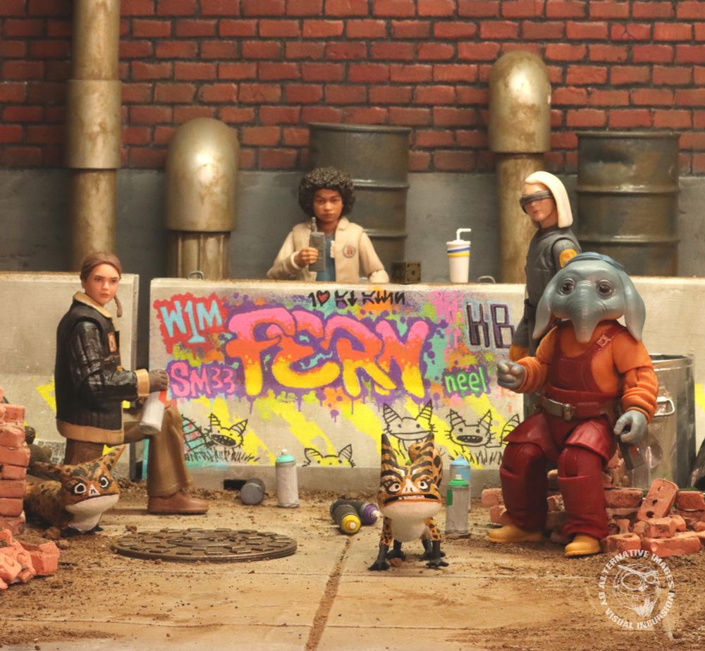 The kids from Skeleton Crew have tagged a concrete barrier and drawn Loth Cats in some street art. They are facing the viewer as they have been caught in the act. Fern in kneeling in front of the art with a spray can in her hand. Wim has his walkie talkie in his hand and KB and Neel are looking on. 