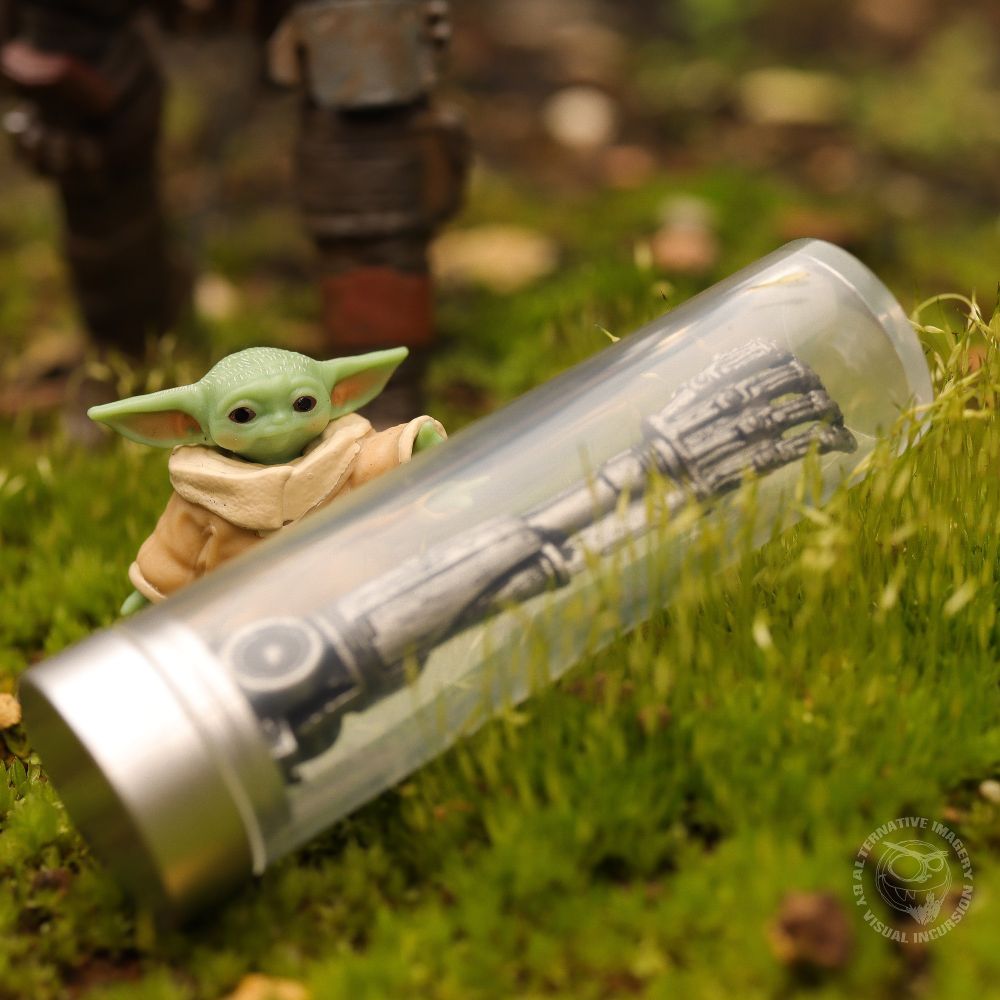 Grogu has found the T-800 arm whilst Mandalorian walks up behind him.  The arm is in the glass display case lying in the grass. Grogu is touching the glass. 