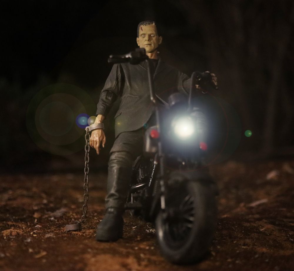 Frankenstein's monster sits on a motorbike with a chain binder hanging from his arm. 