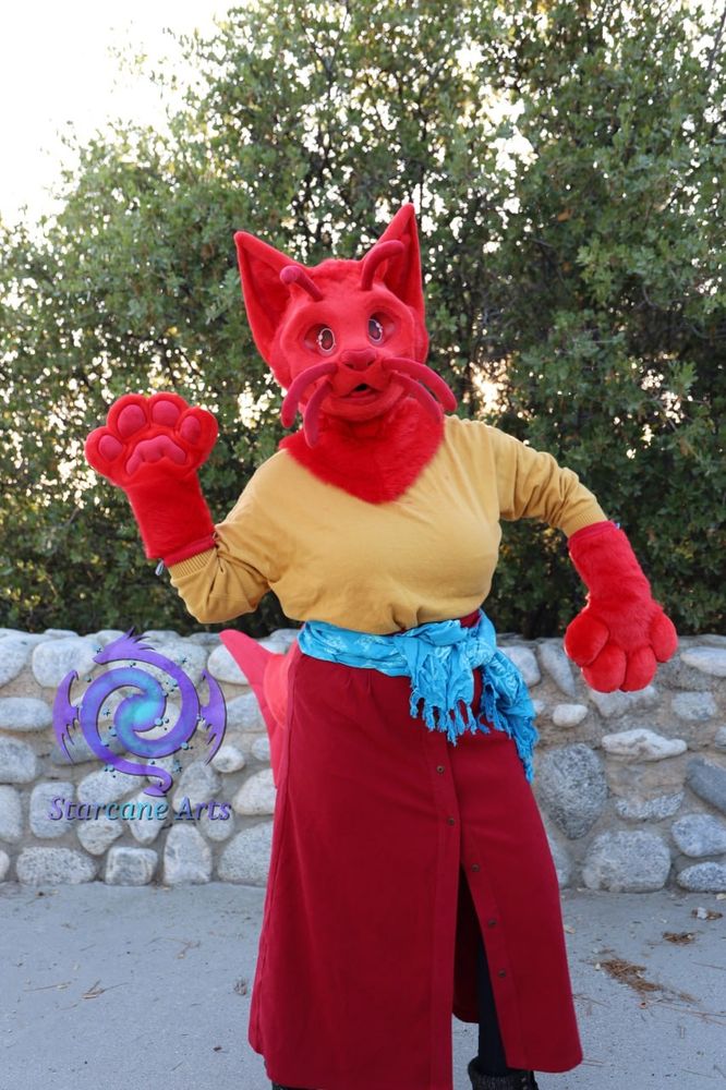 A bright red "cat-fish" fursuiter in a forest environment.